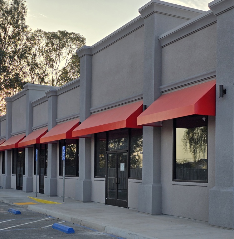 Smooth grey commercial stucco finish on a multi-unit retail building featuring architectural pilasters and vibrant red awnings.